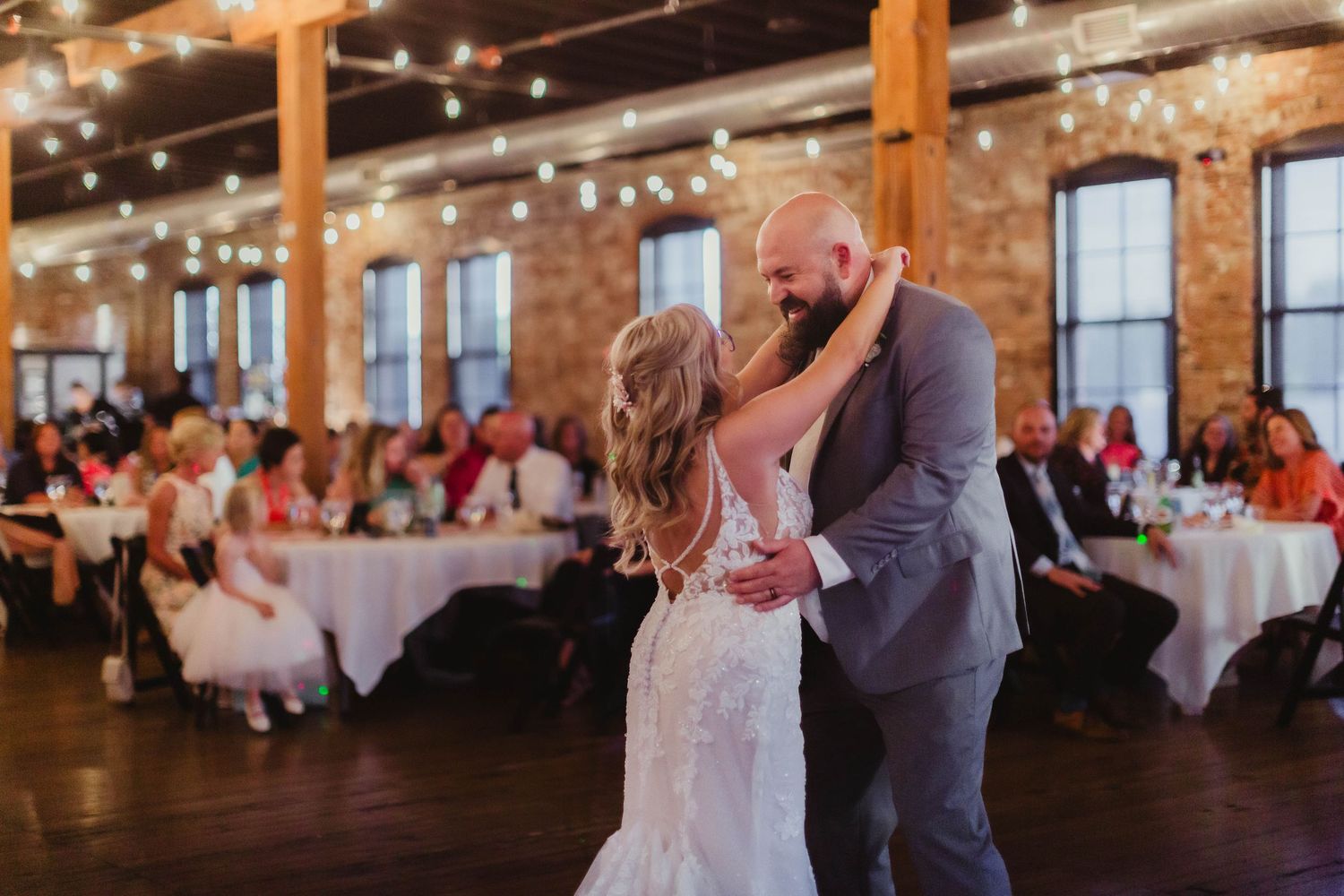 Newlyweds sharing intimate first dance under string lights in rustic venue.