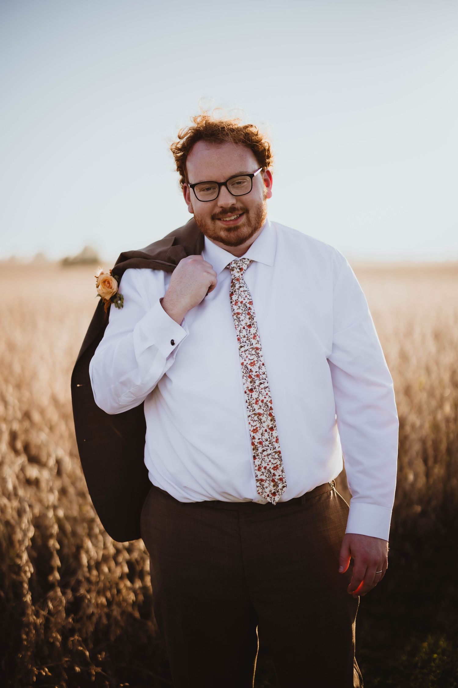 Groom in floral tie and dress shirt posed in front of autumn field at sunset.