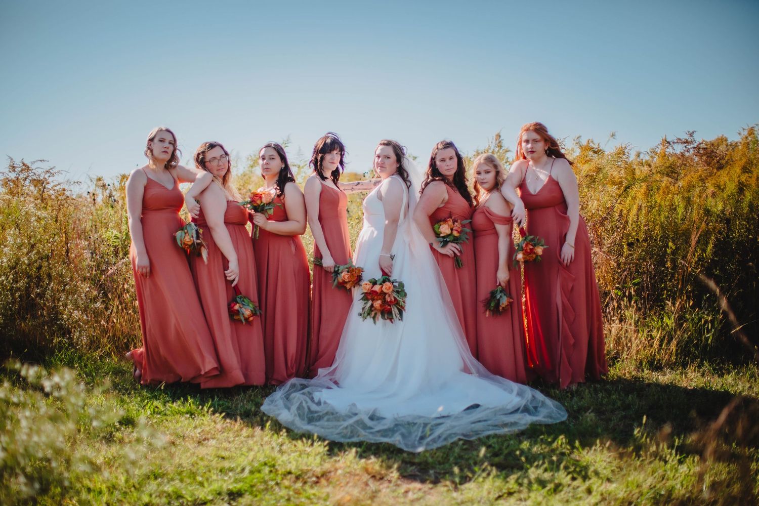 Wedding party in coral dresses arranged for formal photo with bride in white gown outdoors.