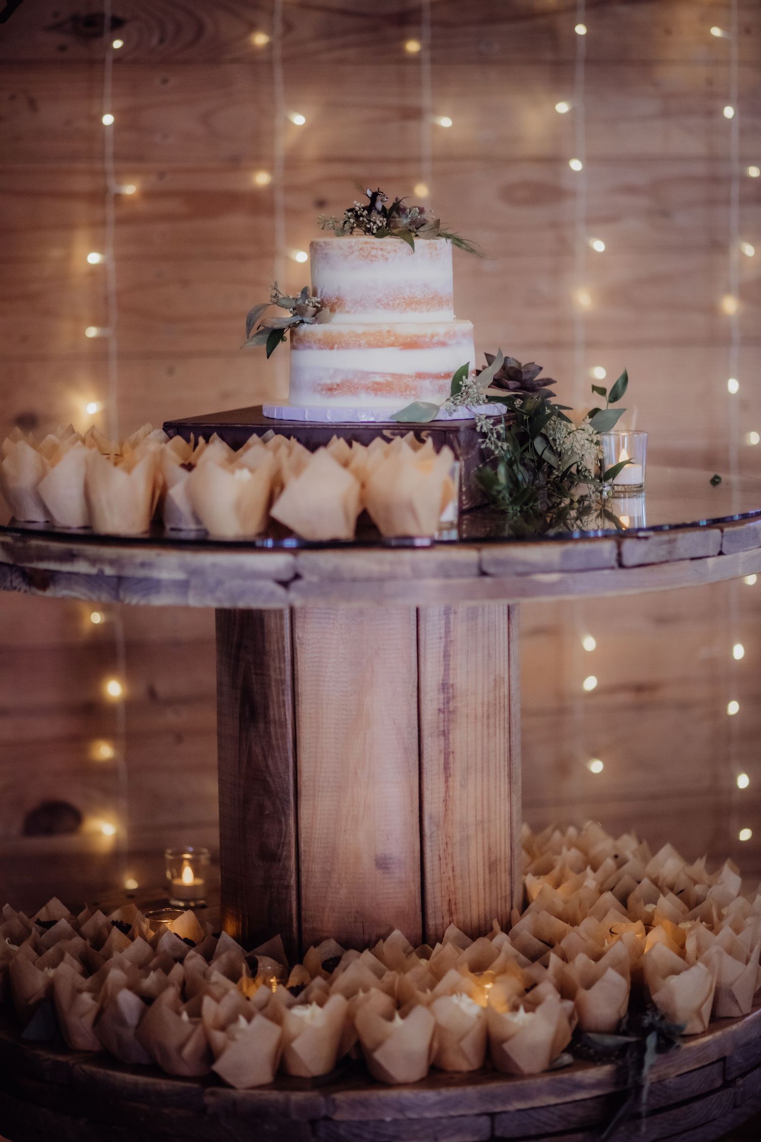 Rustic wedding cake display with white frosted tiers and greenery on wooden stand surrounded by fairy lights.