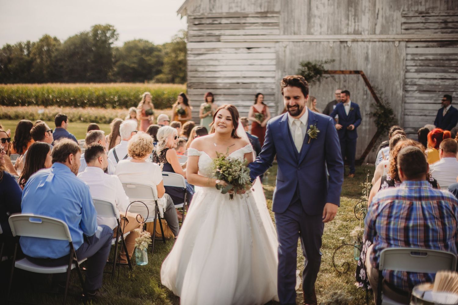 Wedding couple walking down aisle between seated guests at rustic barn venue.