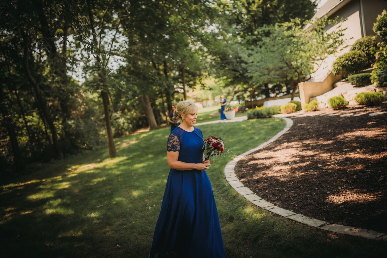 Bridesmaid walks along curved garden path holding floral bouquet.