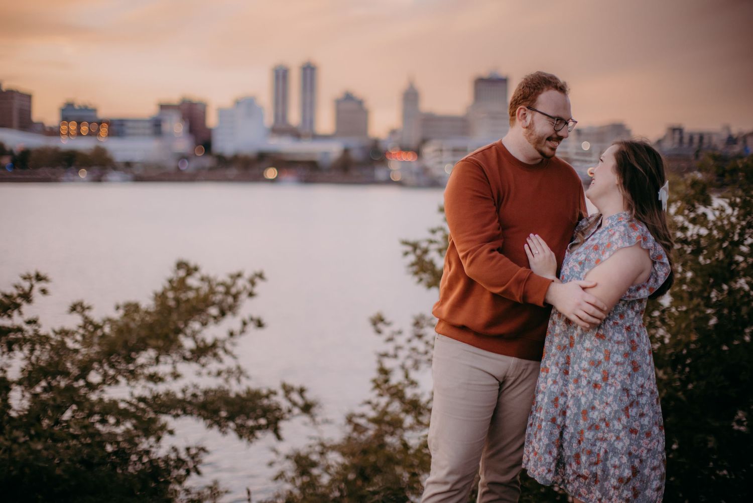 A romantic sunset photoshoot by the water with city buildings creating a dramatic backdrop.