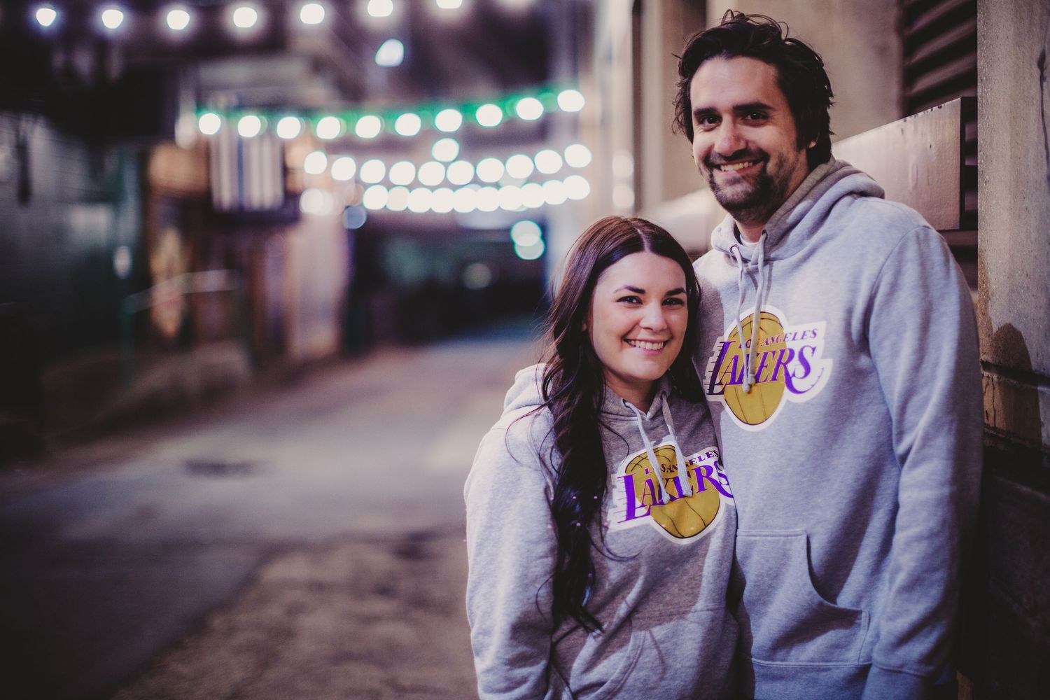 Couple wearing matching Lakers hoodies standing together under string lights at night.