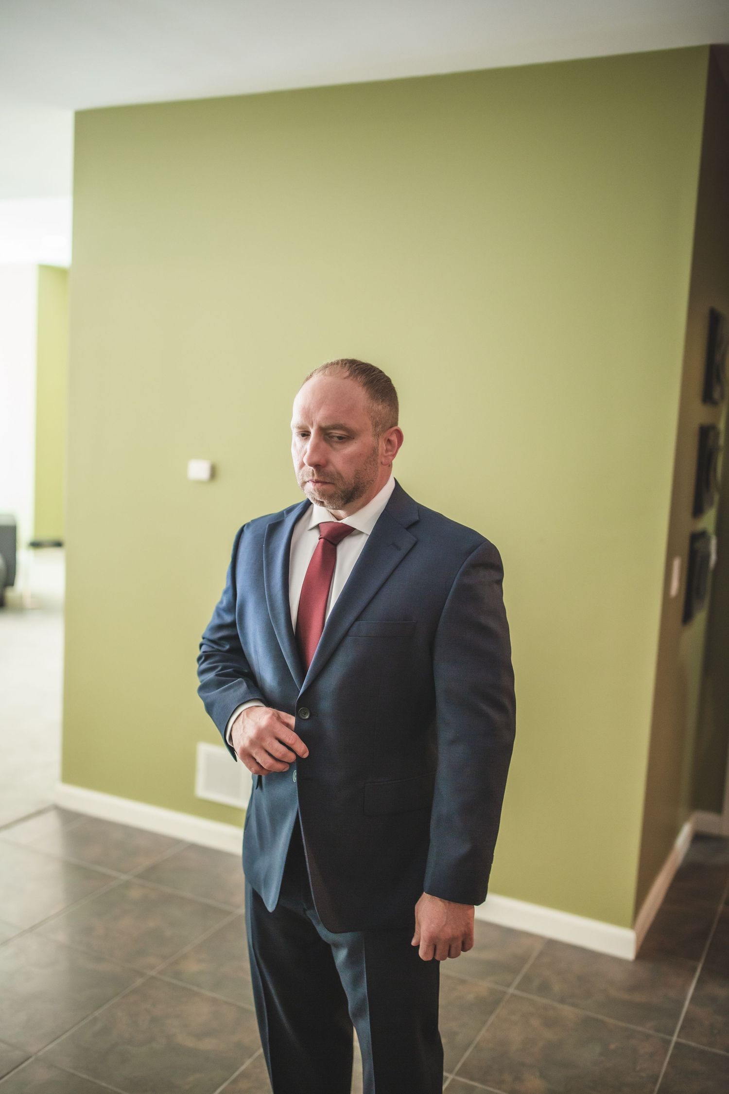 A man in a navy suit and red tie stands in formal attire against a sage green wall.