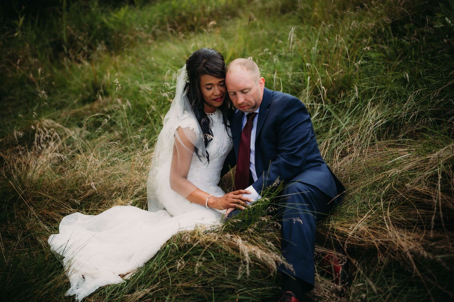 Candid wedding photo of couple sitting in tall grass with natural sunlight filtering through trees.