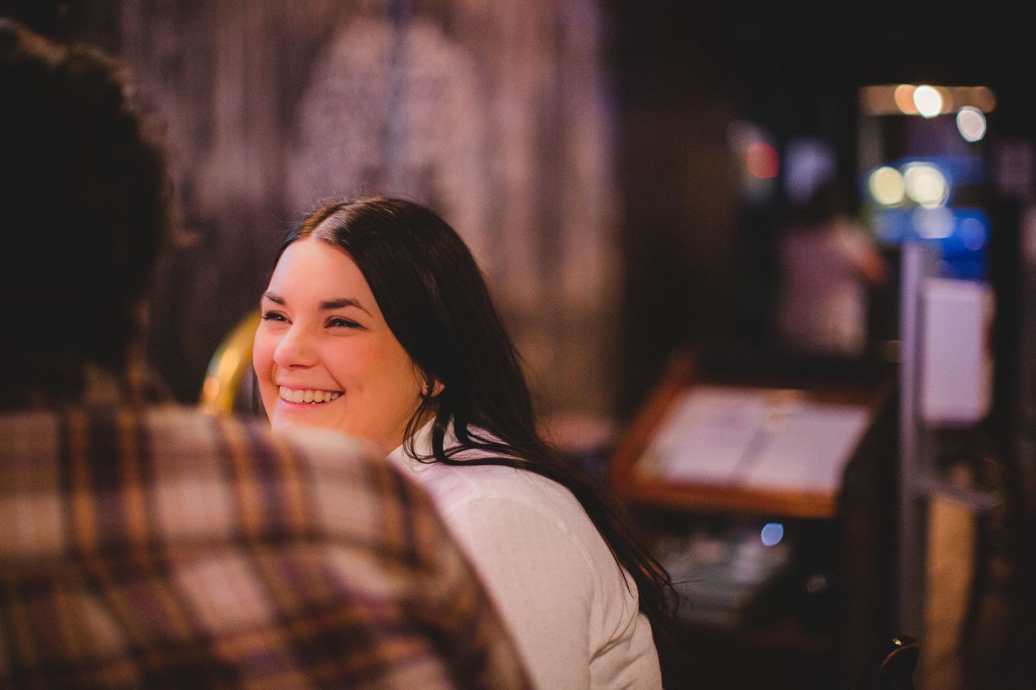 Woman smiling brightly in a cozy bar environment with warm ambient lighting.