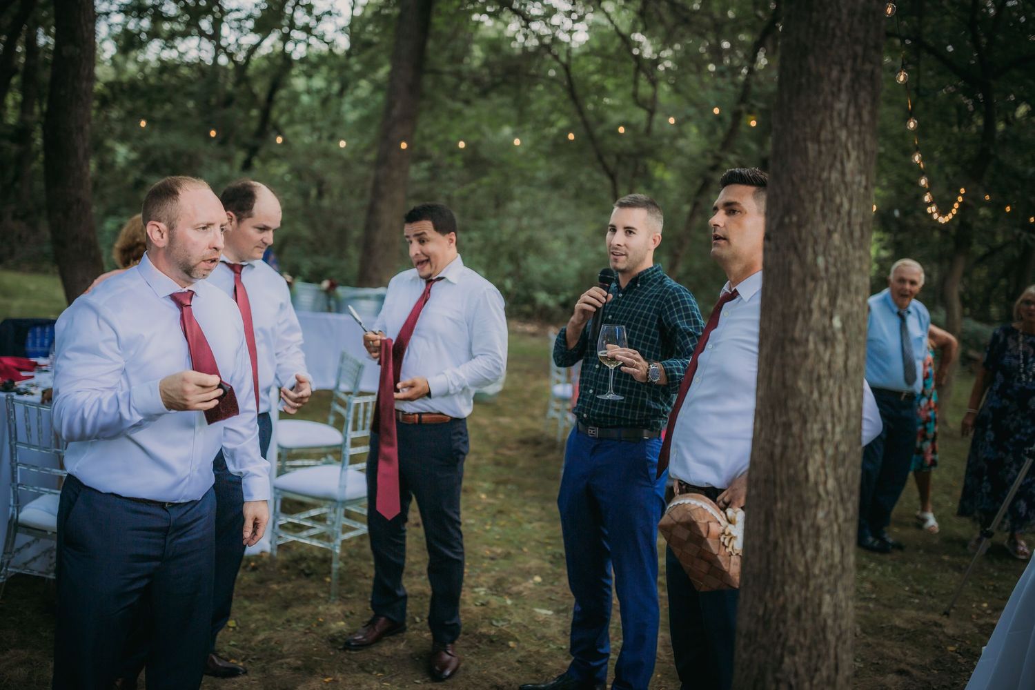 Group of men in dress shirts and ties conversing at outdoor wedding reception.