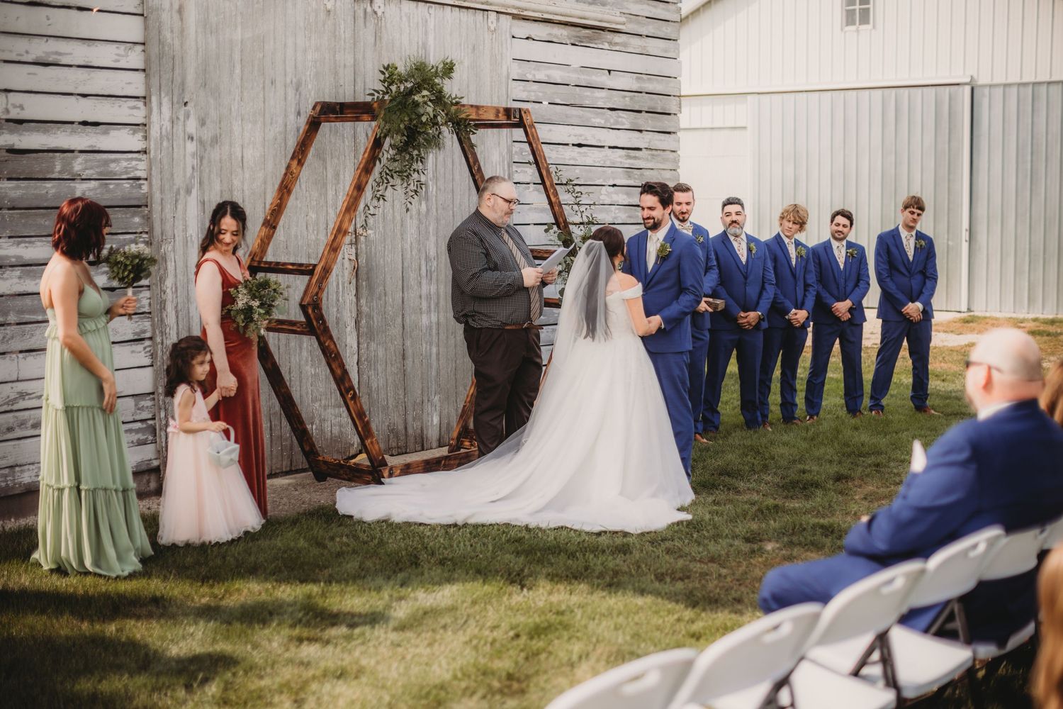 Rustic outdoor wedding ceremony with wooden triangle arch and white barn backdrop.
