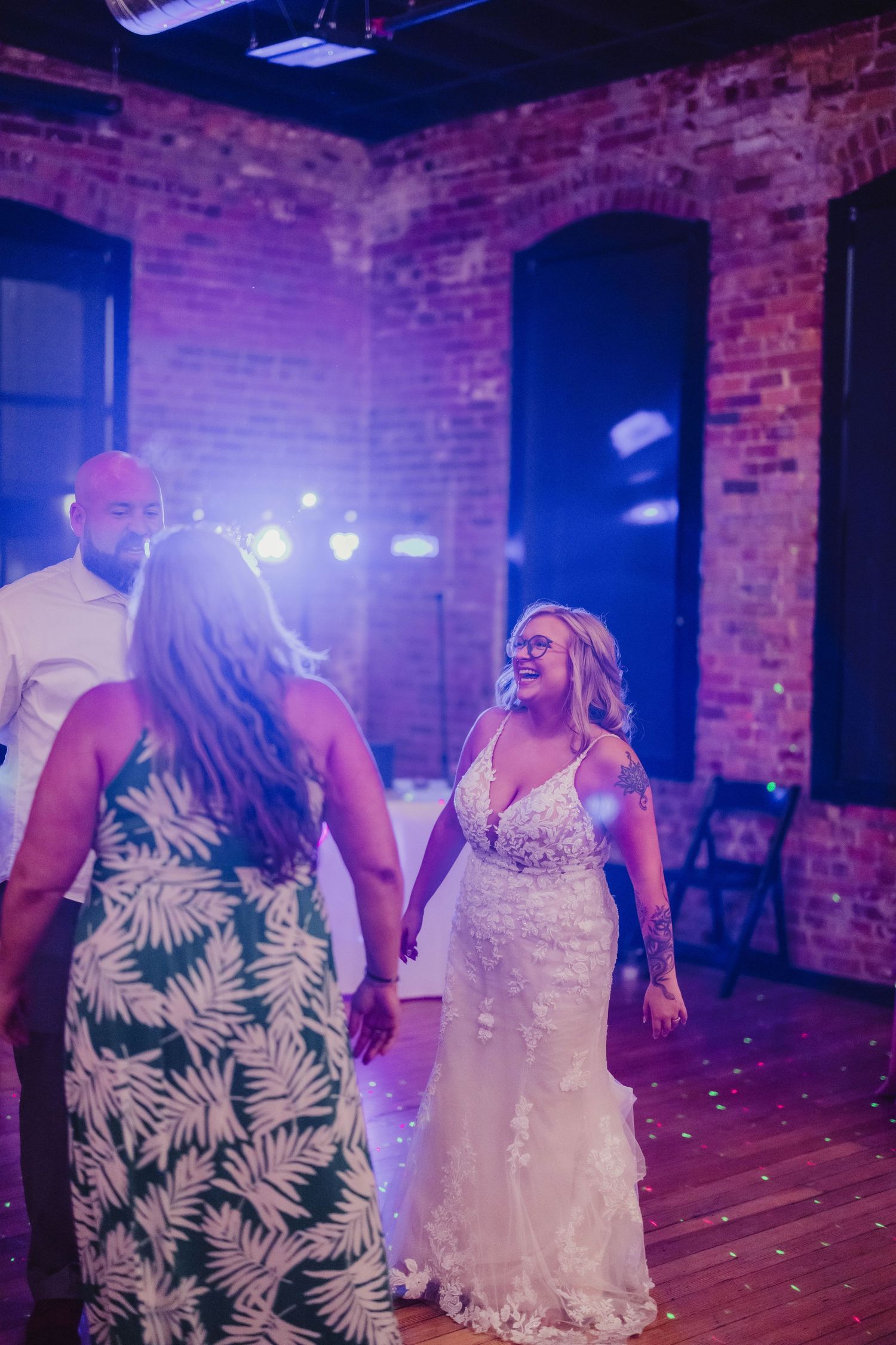 People dancing at a wedding reception in a rustic brick-walled venue with festive lights and decorative palm print dresses.