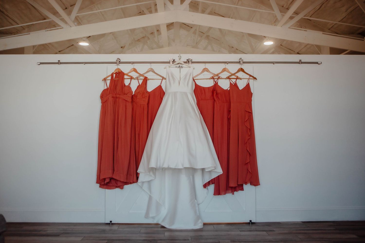 White wedding dress hanging between coral bridesmaid dresses under vaulted ceiling.
