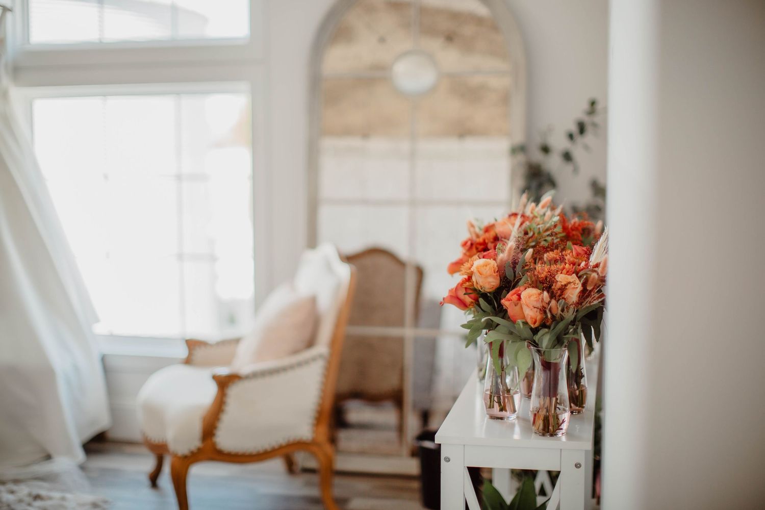 Elegant sitting area with arched window and coral floral arrangements on white furniture.