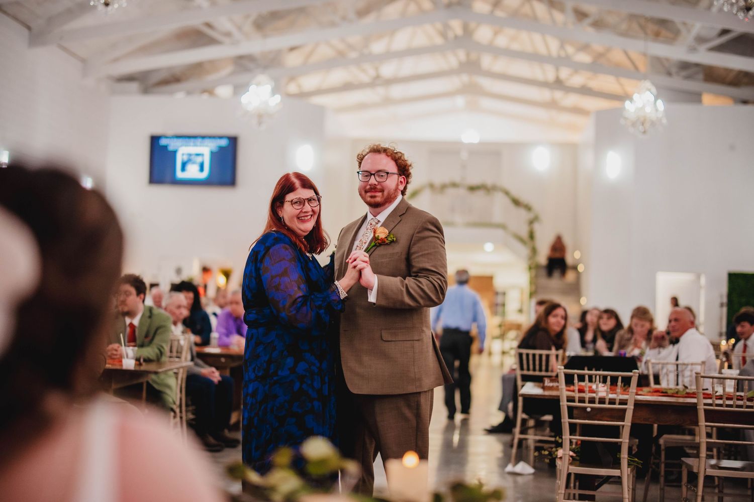 Two people dancing together at an indoor event with exposed beam ceilings and chiavari chairs.