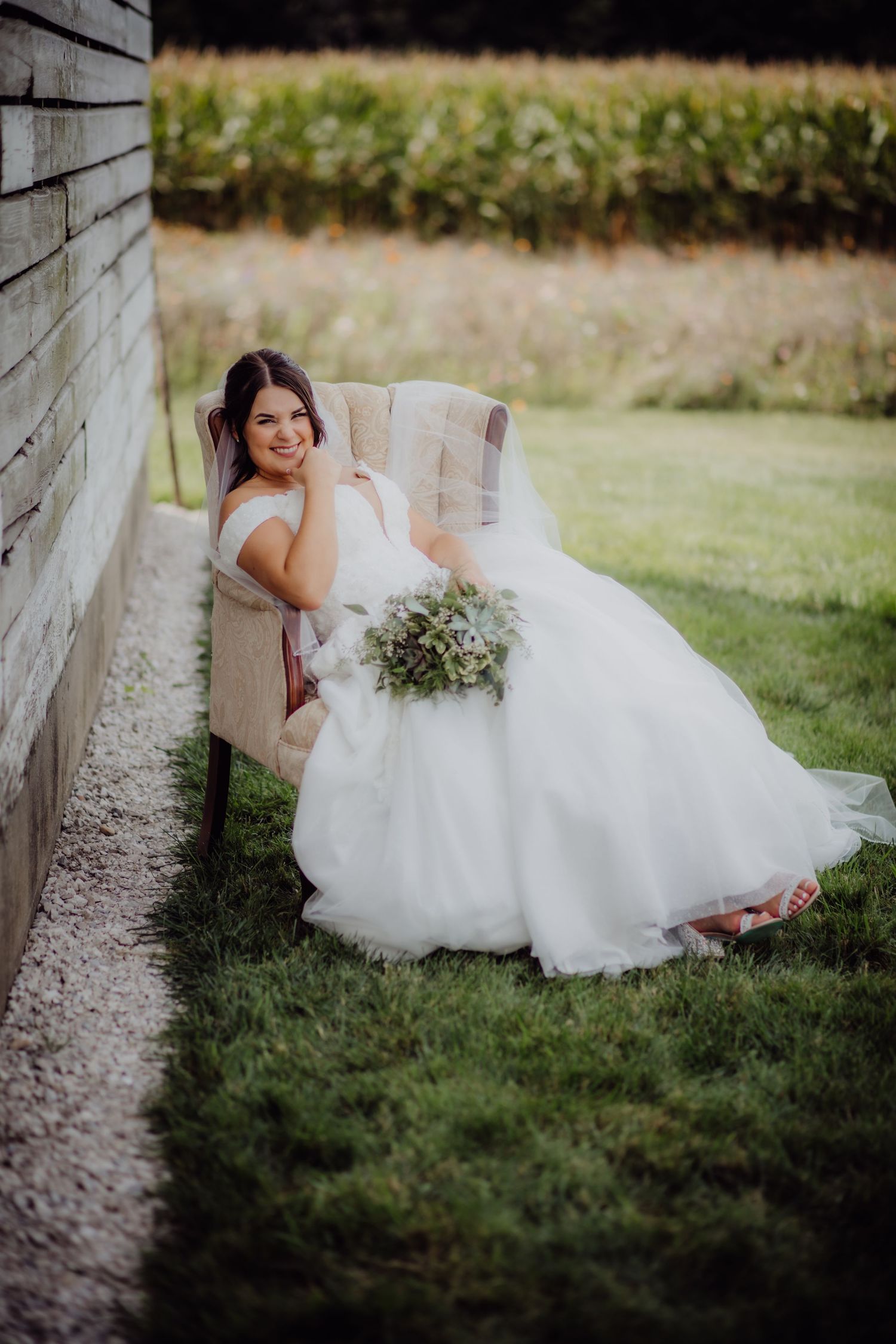 Beautiful wedding dress with flowy tulle skirt and delicate bouquet photographed against rustic barn wall and green lawn.