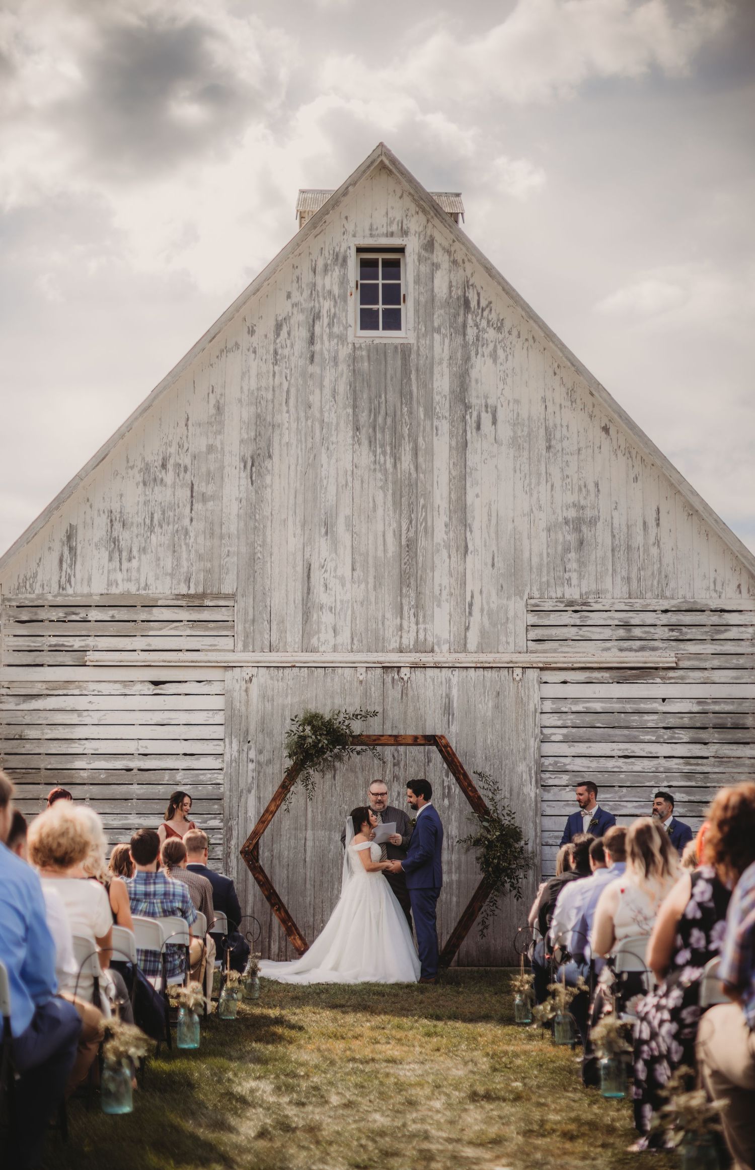 A rustic farmhouse wedding ceremony setting features a white barn and scattered chairs on green grass.