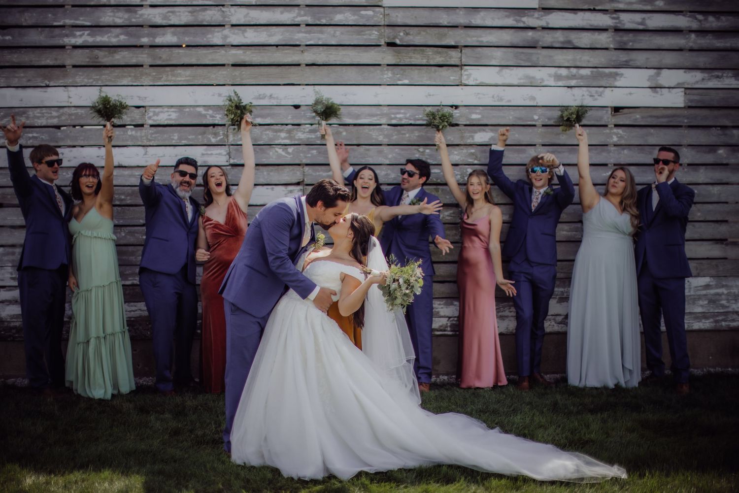 Wedding group stands in formation wearing coordinated purple and pastel dresses against barn wall.