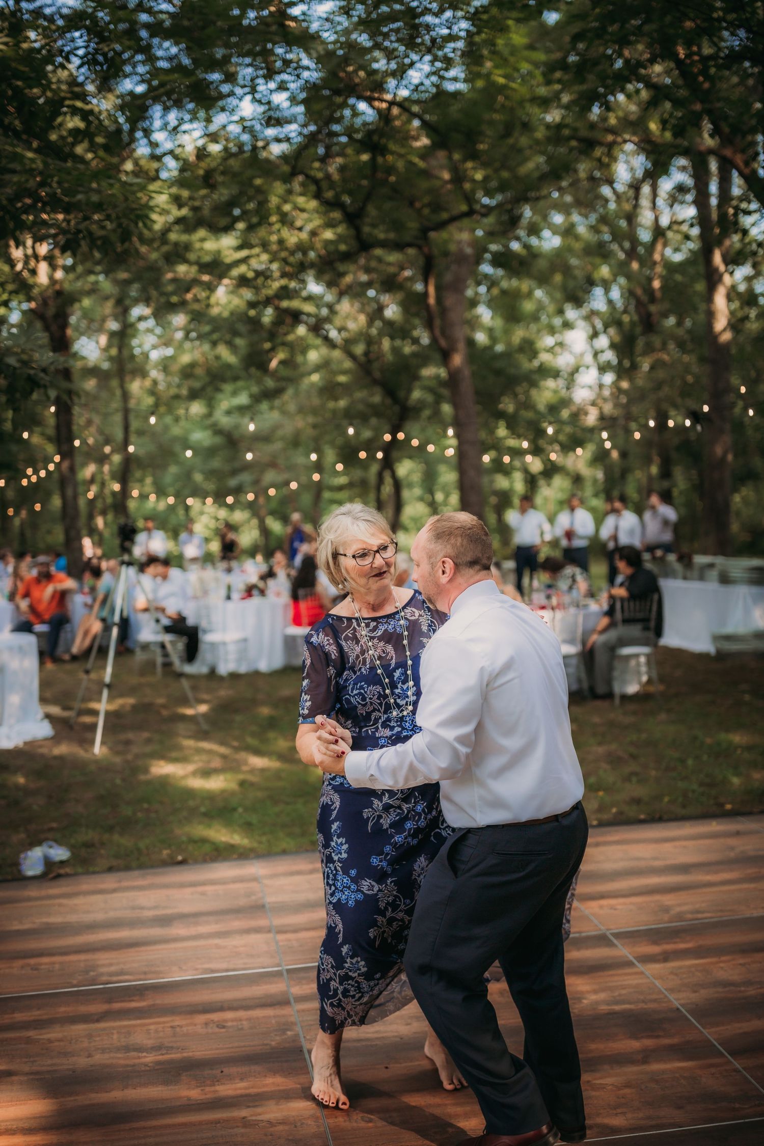 Couples share an intimate dance moment on wooden platform at outdoor forest reception with string lights.