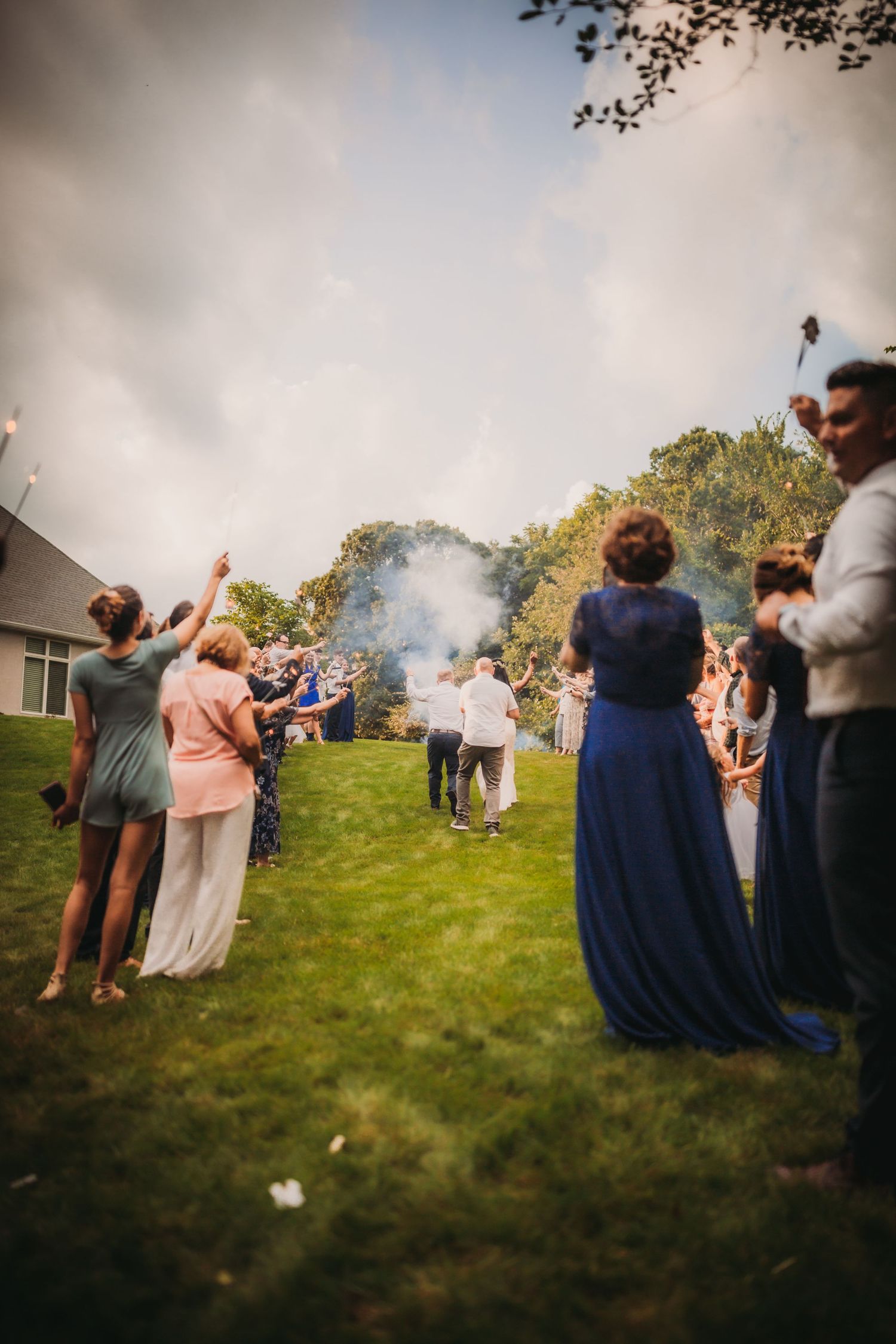 A group watches from a grassy slope during an outdoor wedding celebration with atmospheric smoke.
