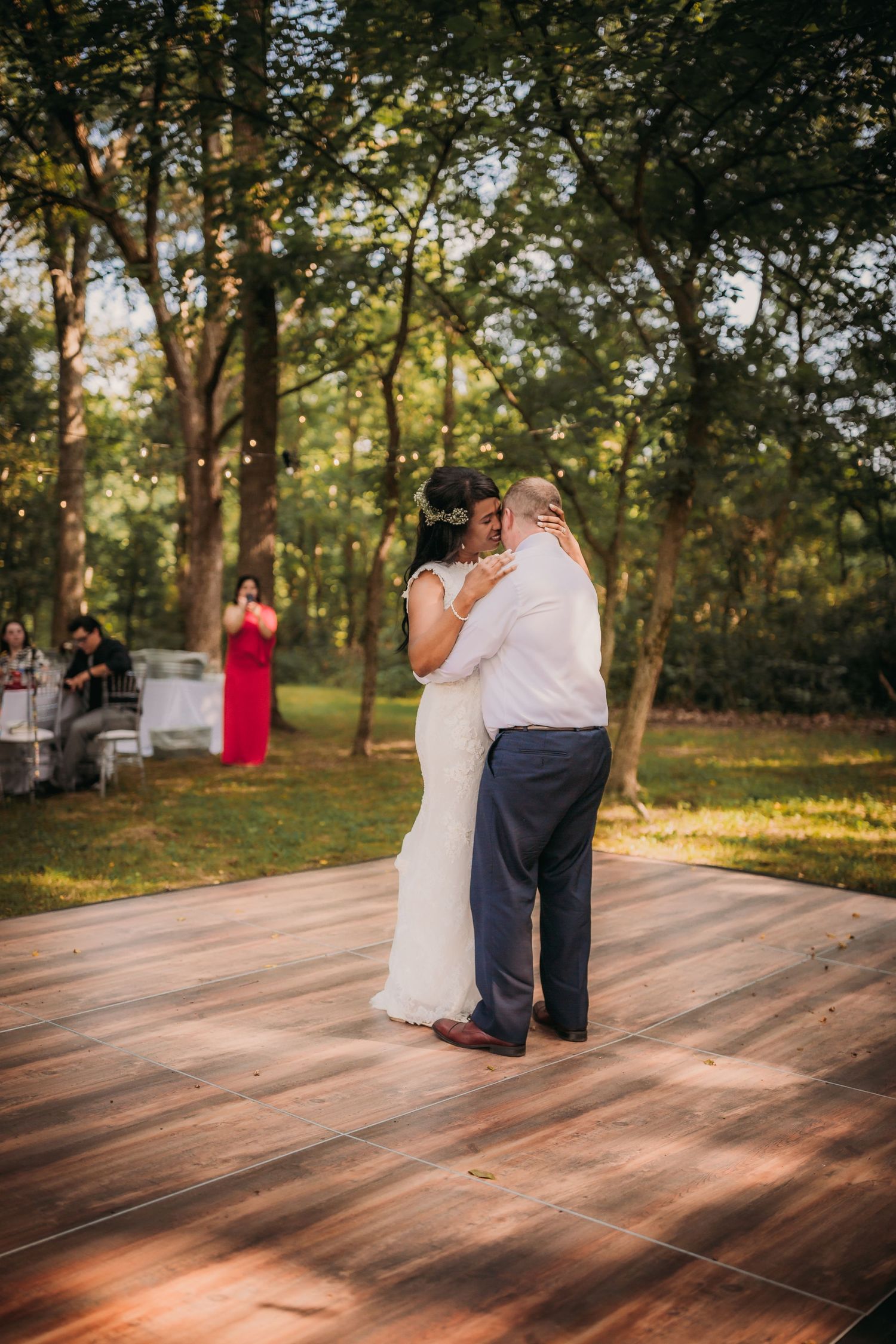 Newlyweds share a romantic dance moment on an outdoor wooden platform with trees in the background.
