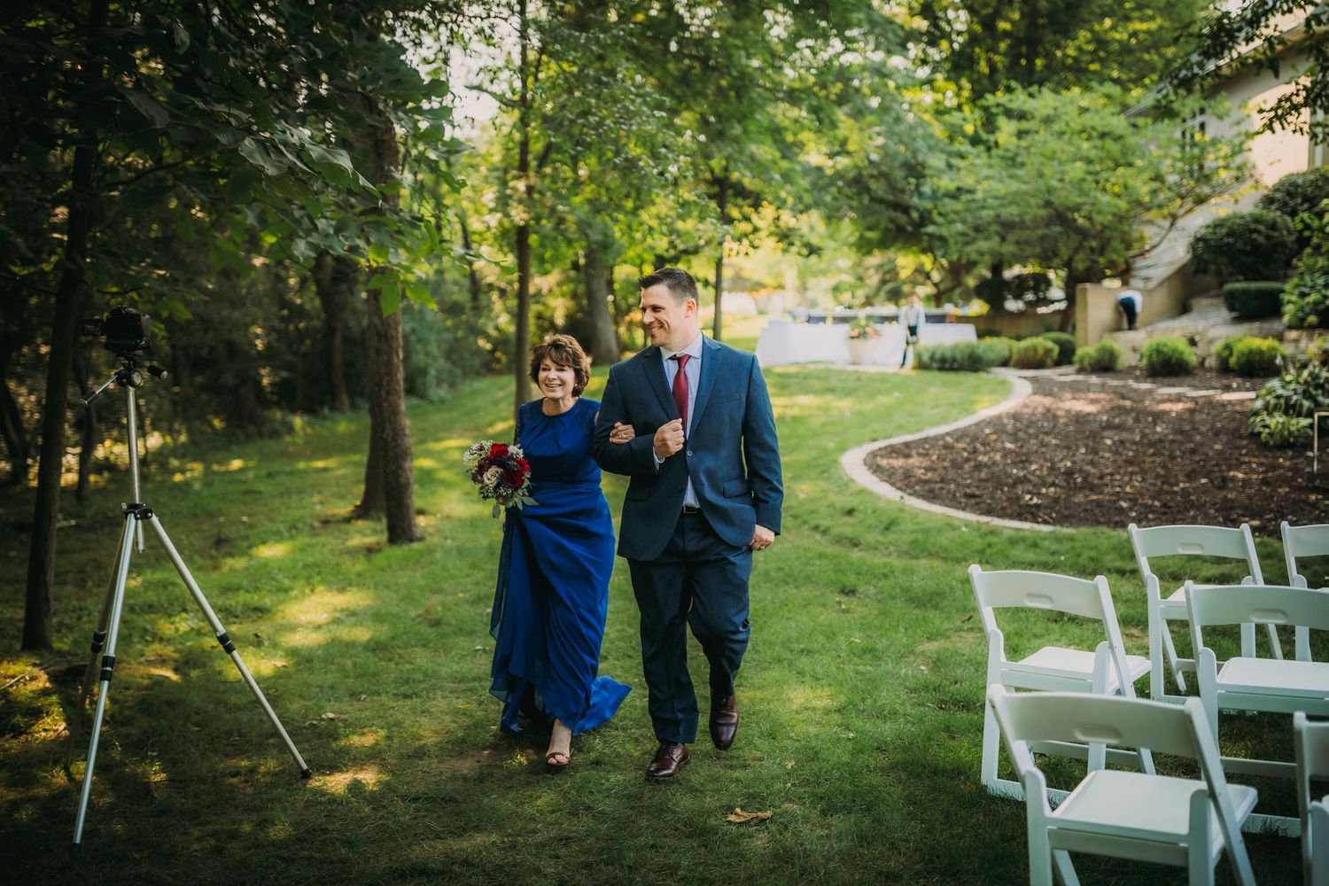 Wedding attendants make their way down a sun-dappled garden ceremony aisle.