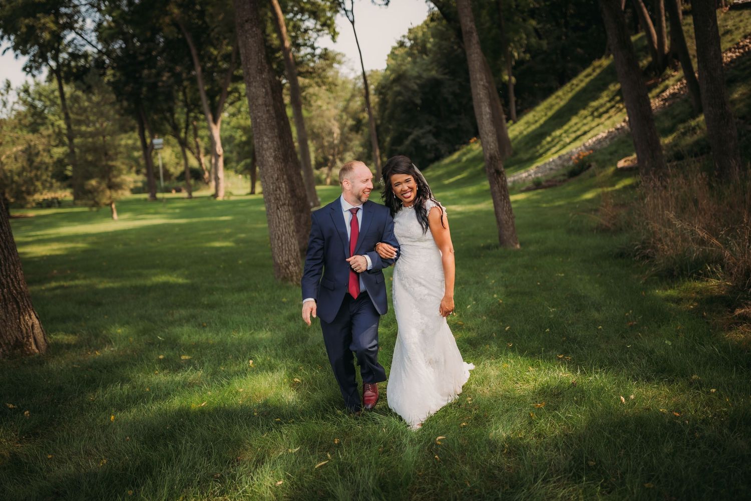 Newlyweds walk together on a grassy hill surrounded by tall trees.