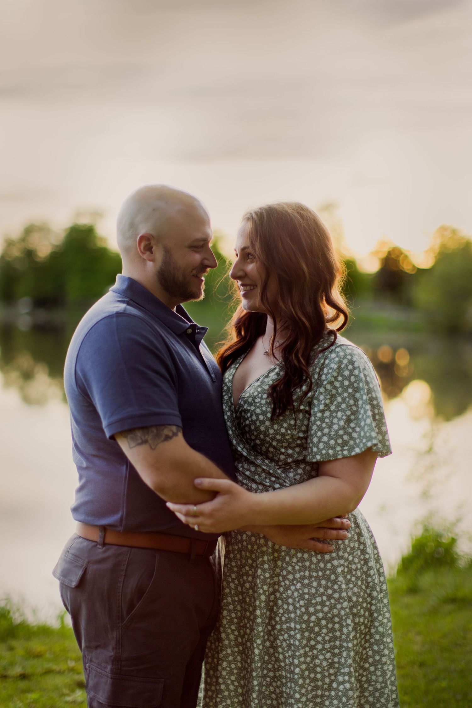 A romantic outdoor photoshoot at sunset with a couple embracing on grassy lakeside grounds.