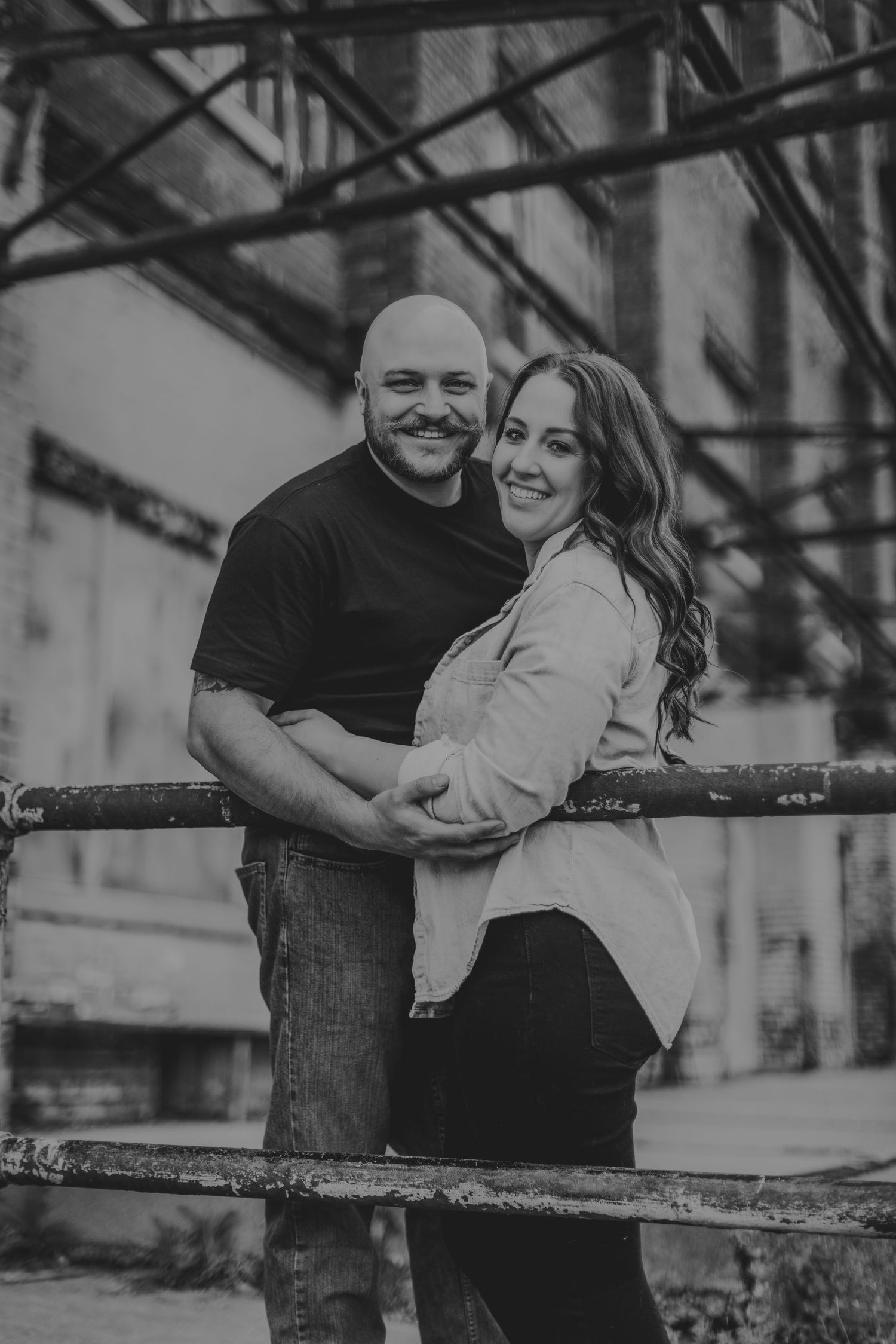A black and white portrait of a couple hugging against an urban industrial backdrop.