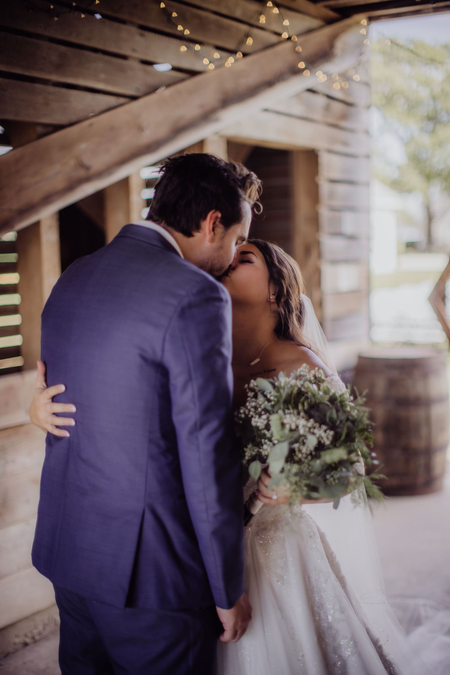A romantic kiss between newlyweds in a rustic barn setting with string lights overhead.