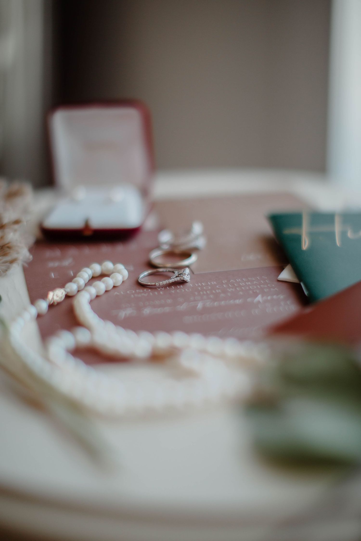 Pearl jewelry and wedding rings displayed with burgundy and teal wedding stationery.