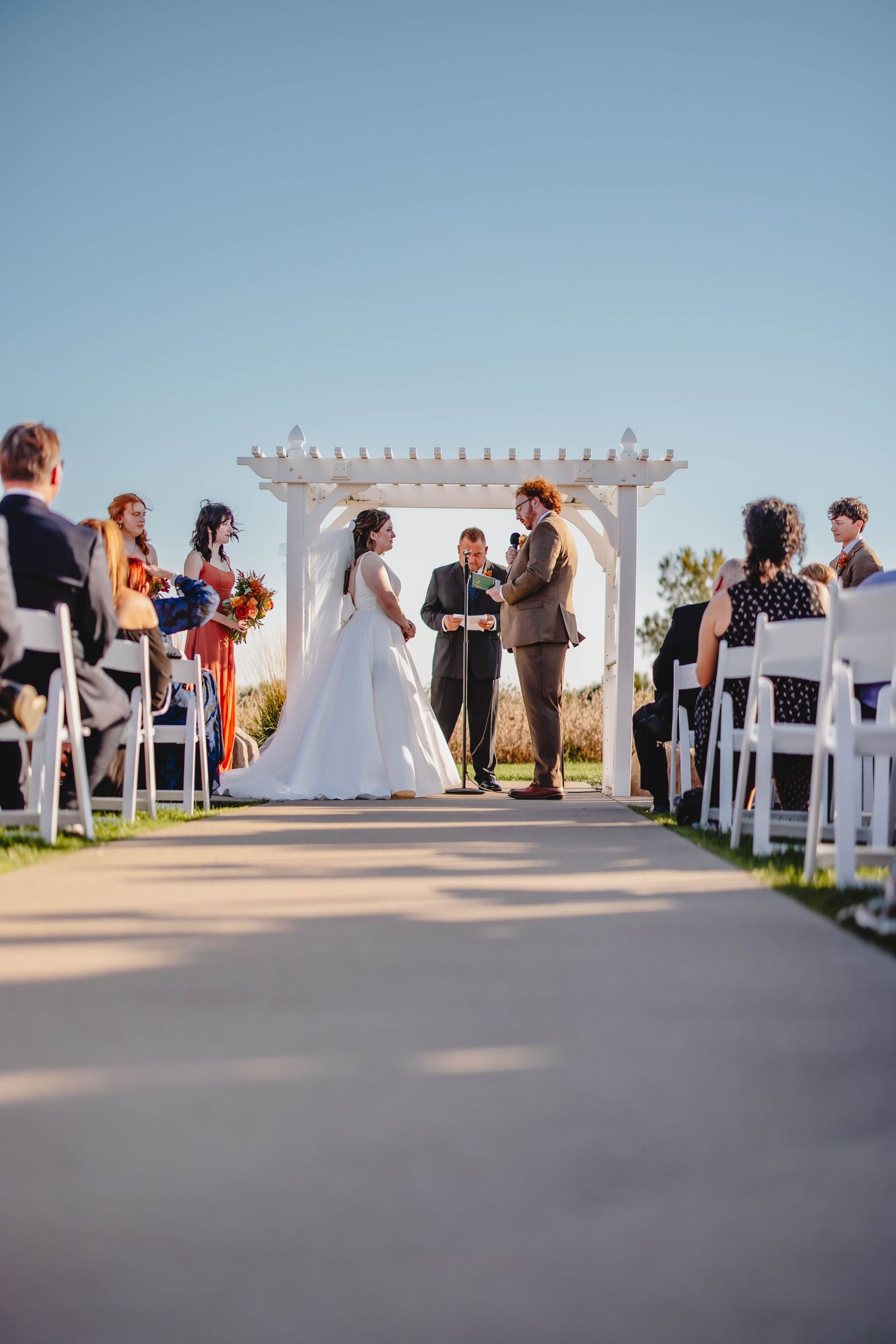 Couple exchanging vows beneath white pergola during outdoor autumn wedding ceremony.