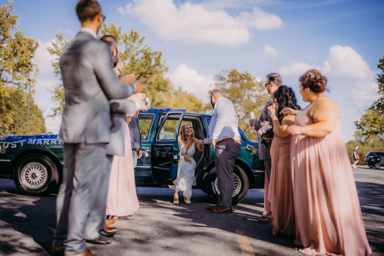 Bridal party celebrating around vintage car with bridesmaids in pink dresses and groomsmen in gray suits.