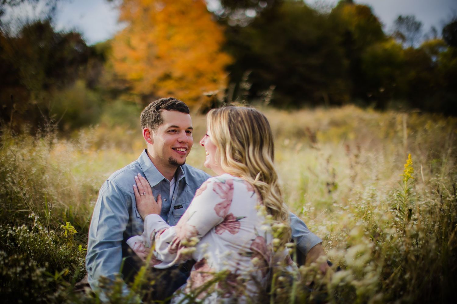 Romantic couple sharing a laughing moment while sitting in tall grass during sunset with fall foliage behind them.