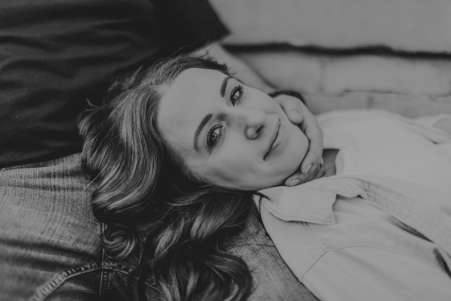 A close-up black and white portrait of someone with wavy hair lying down and smiling.
