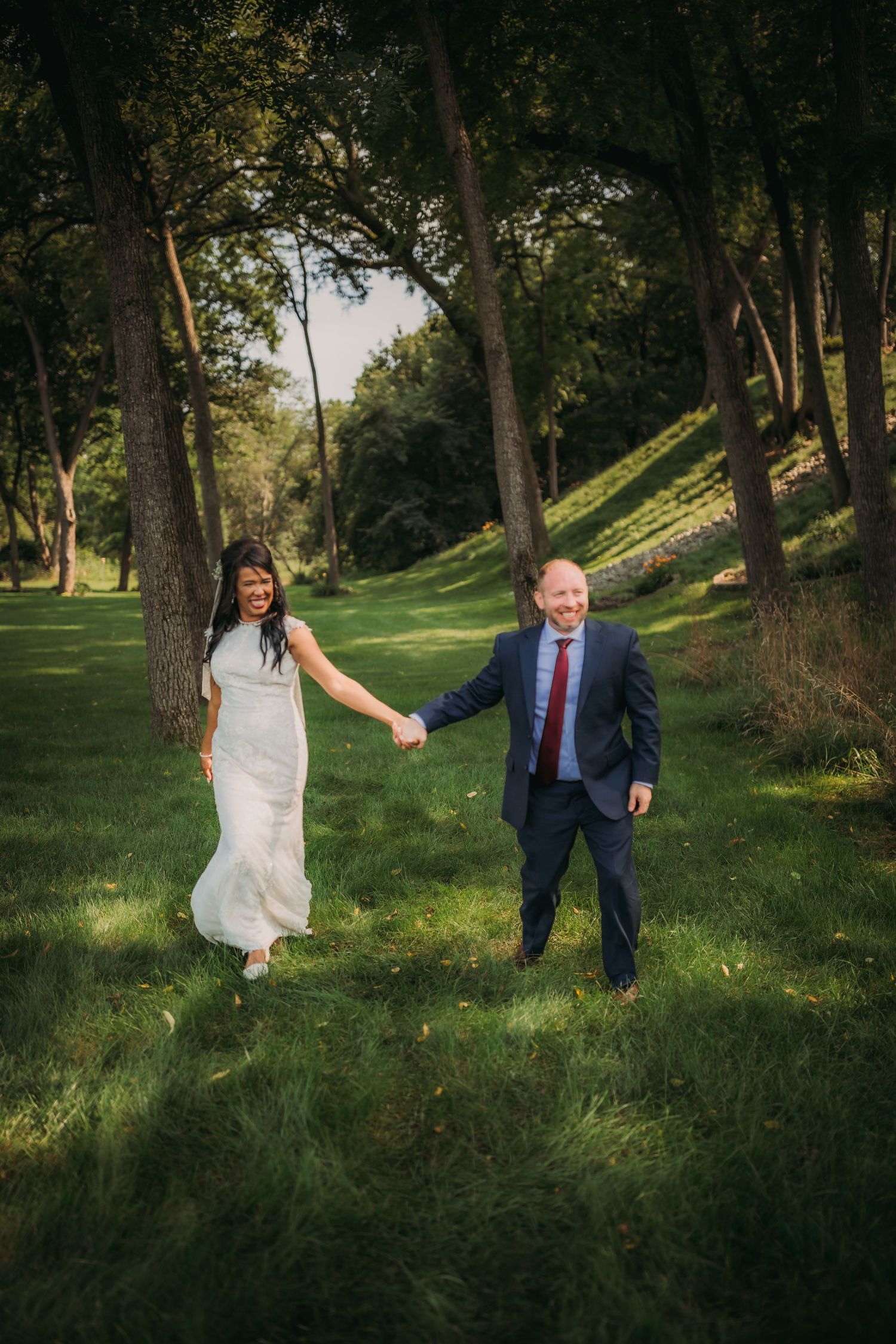 A bride and groom hold hands while walking through a sunlit forest glade.