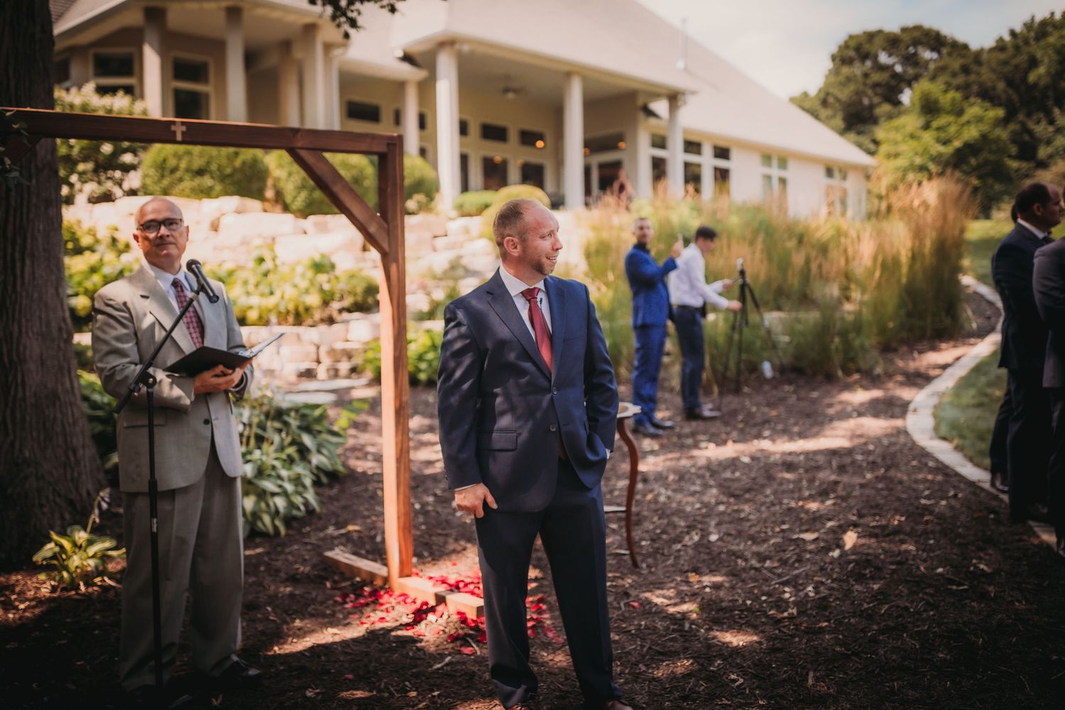 People stand near a wooden arch outside a colonial-style building before a ceremony.