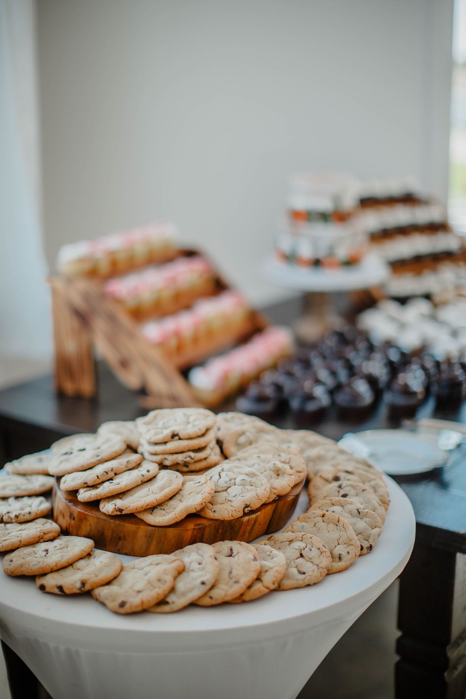Fresh chocolate chip cookies displayed on white serving platters at a dessert table.