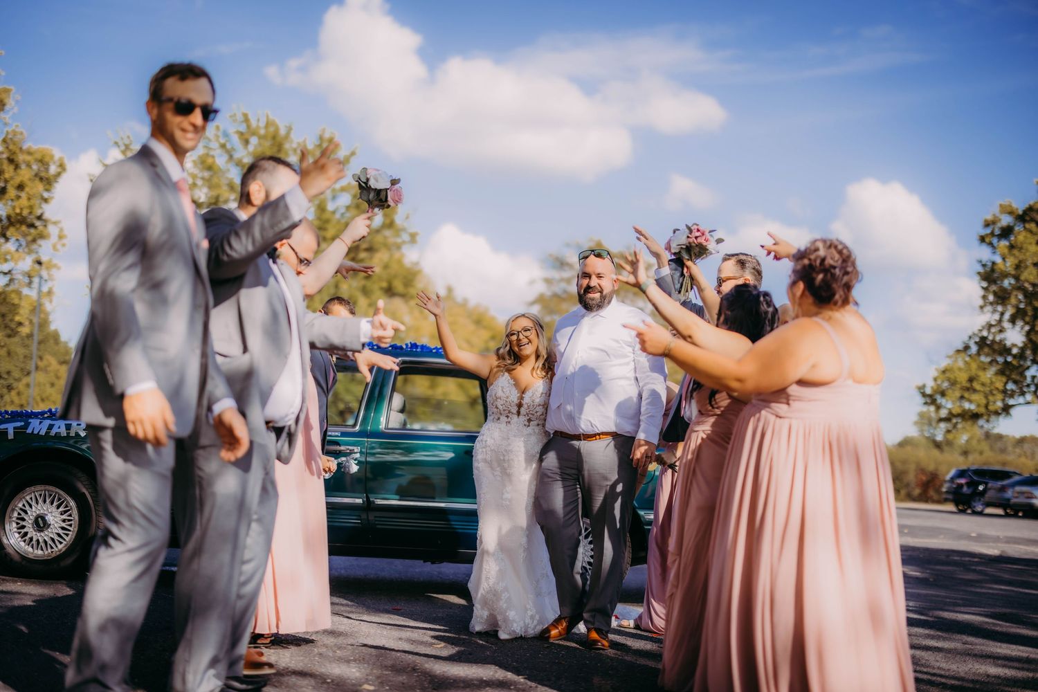 Wedding party making celebratory toasts beside vintage car on sunny afternoon.