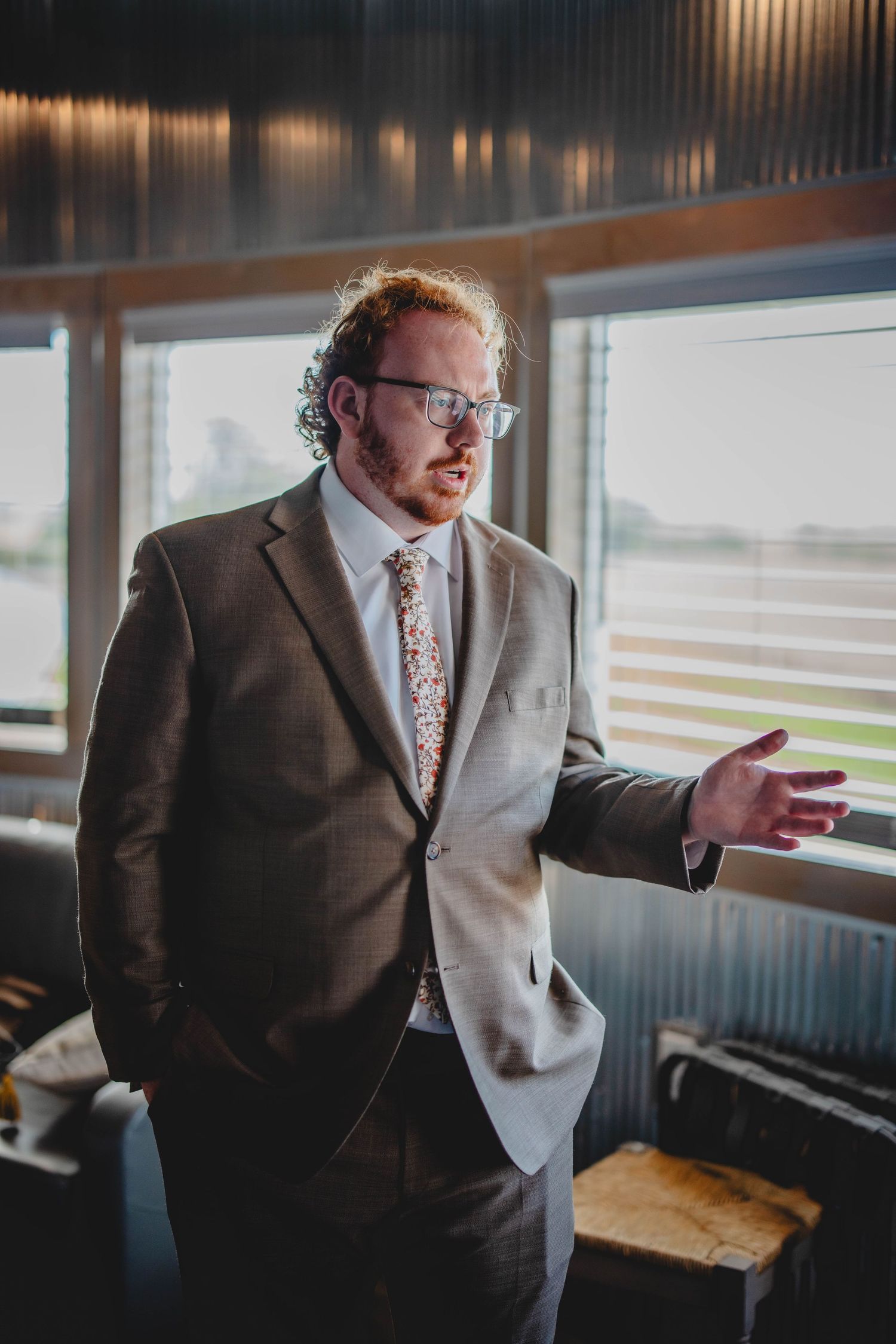 A person in a brown suit and floral tie gazes thoughtfully out a window in a warmly lit room.