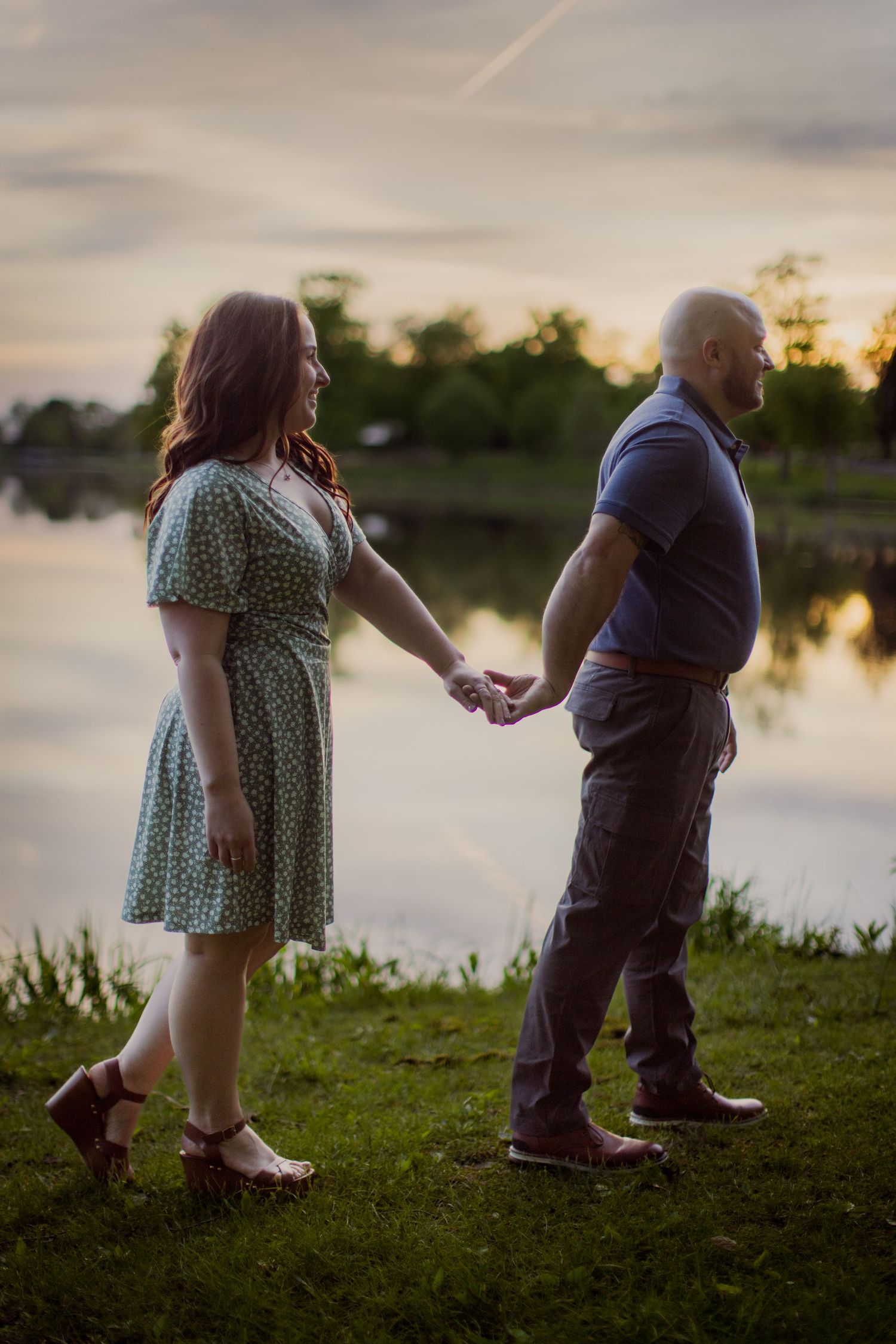 Two people hold hands while walking along the shore of a lake at dusk.