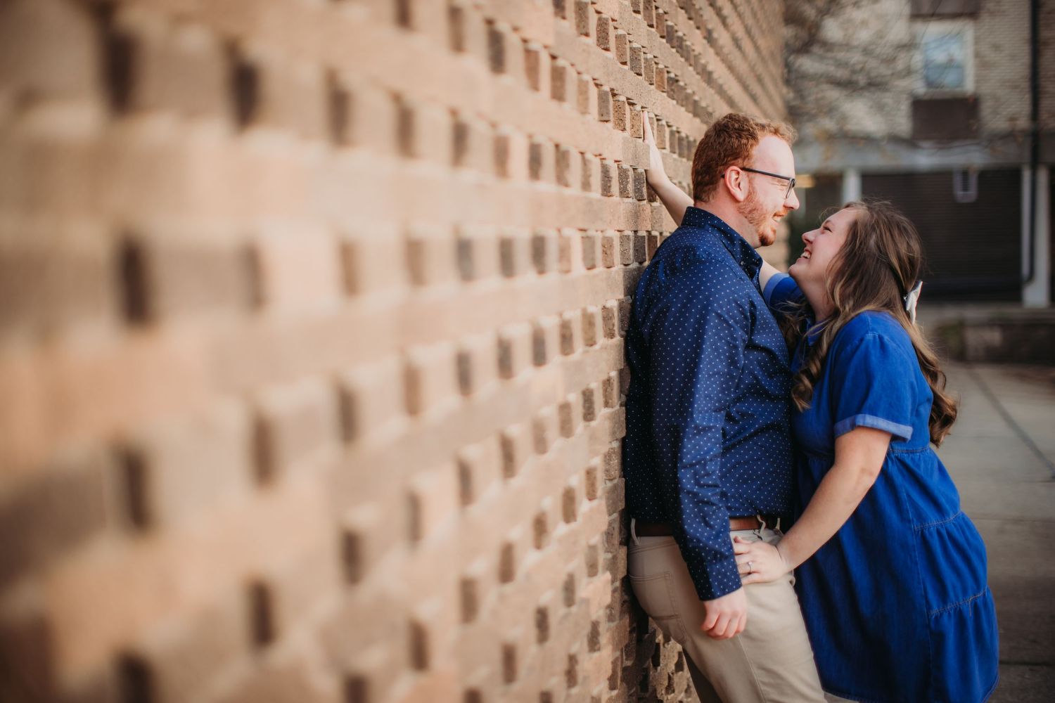 A couple in coordinating blue and khaki outfits share an affectionate moment by a textured wall.