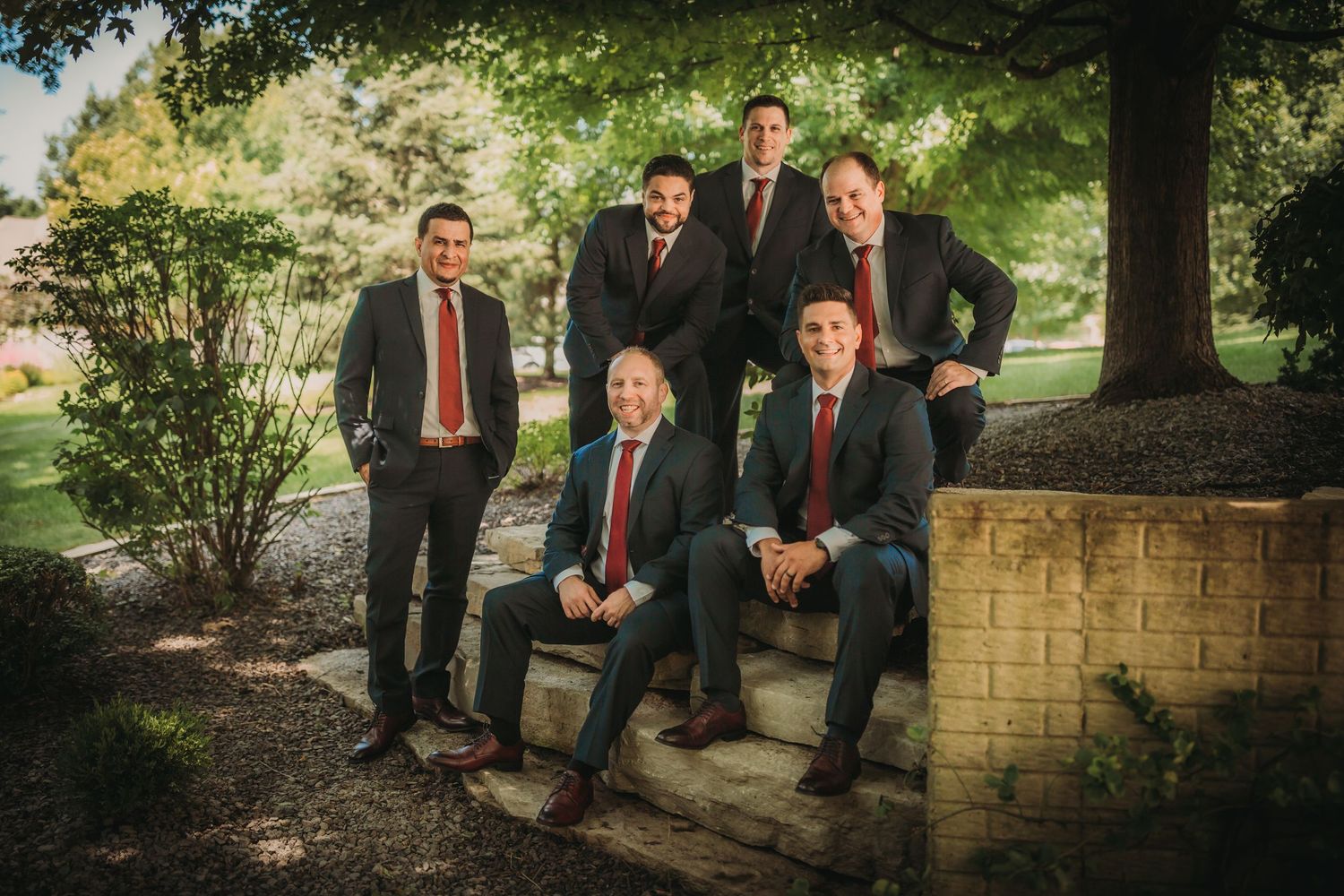 Stylish groomsmen group arranged on stone wall with lush green backdrop.