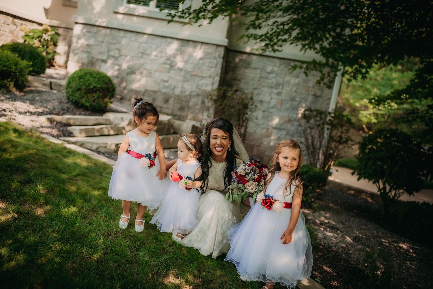 Little attendants in white dresses gather on a garden path with floral bouquets during a wedding celebration.