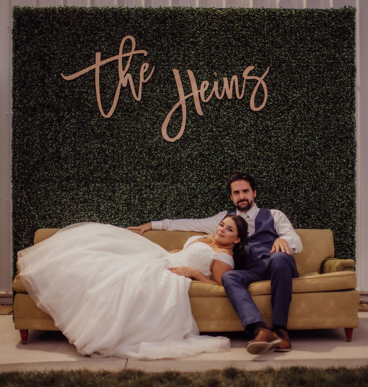 Wedding couple lounging on a tan couch against a dark green hedge wall backdrop.