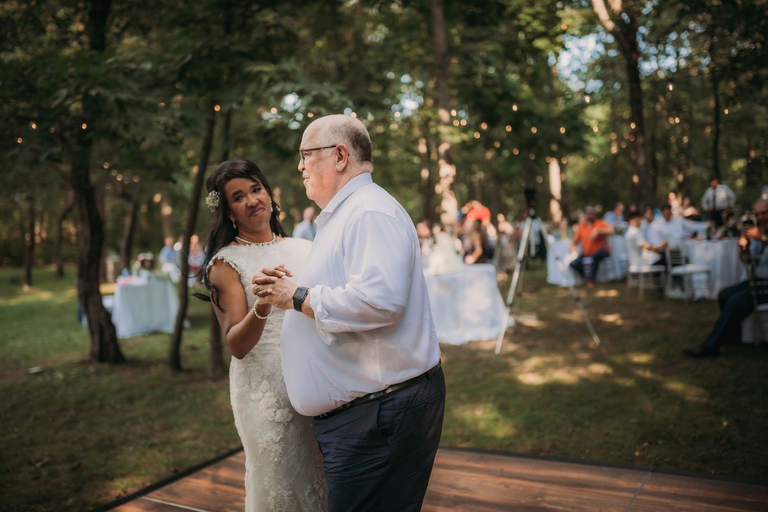 Couples dancing together during outdoor woodland reception with guest tables and string lights visible in background.