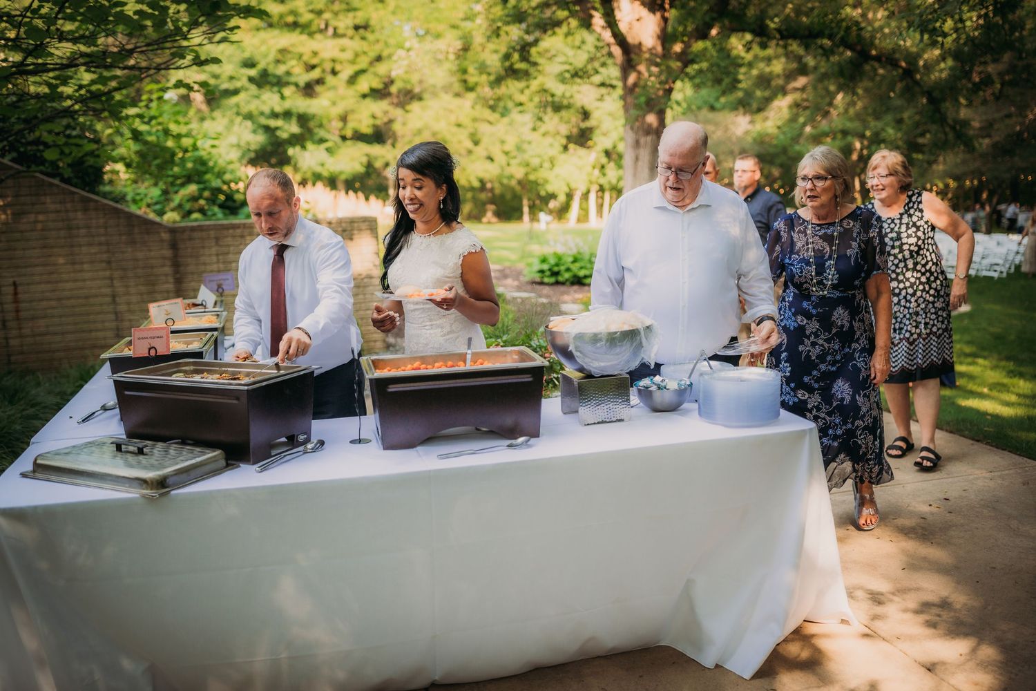 Wedding guests enjoying refreshments at outdoor buffet tables in dappled sunlight.