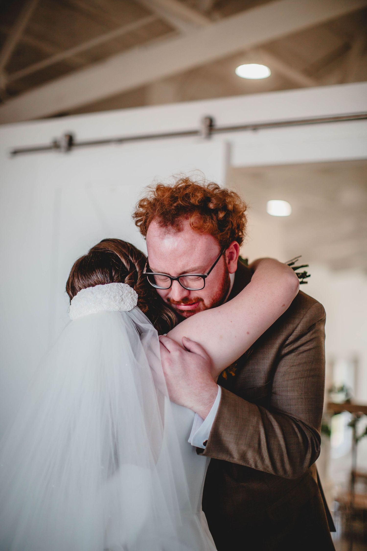 Emotional embrace during wedding first look with white veil and brown suit.