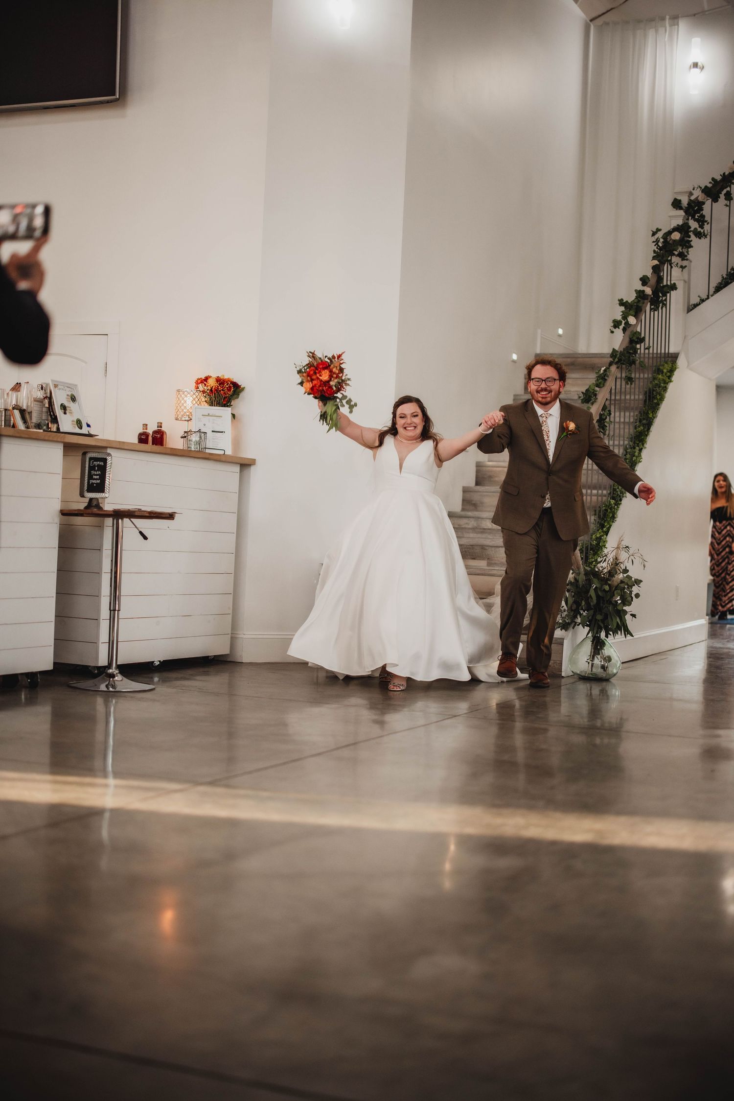 A joyful couple celebrates at their wedding reception walking hand in hand with raised bouquets of orange flowers.