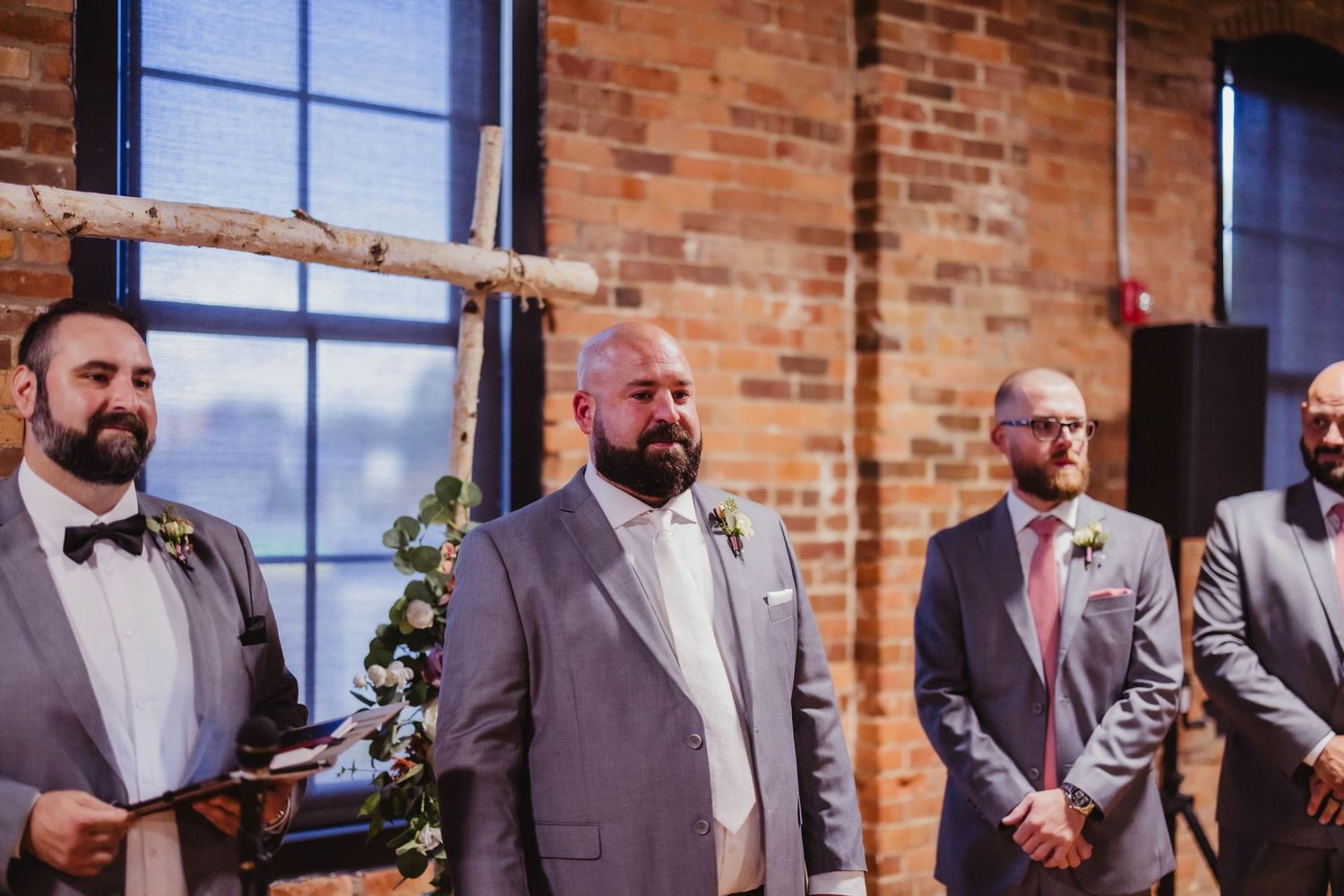 Groomsmen in grey suits stand at the altar during a wedding ceremony in an industrial-style venue.