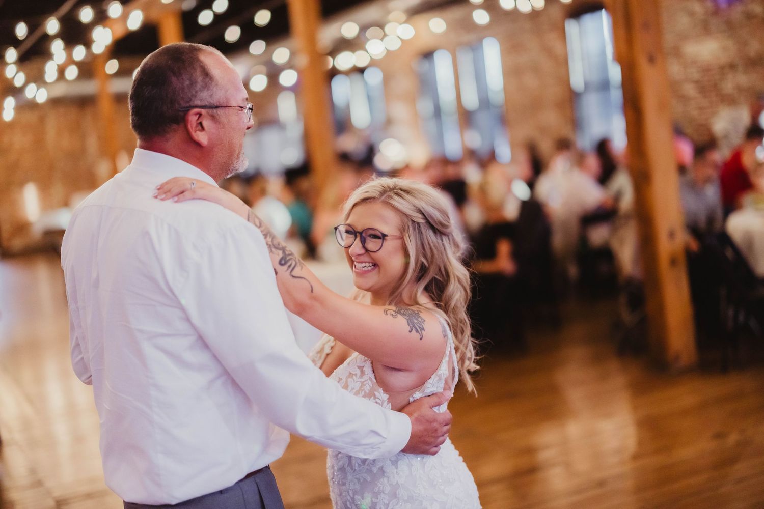 Wedding reception dancers move across a rustic wooden floor under warm string lights in an intimate venue setting.