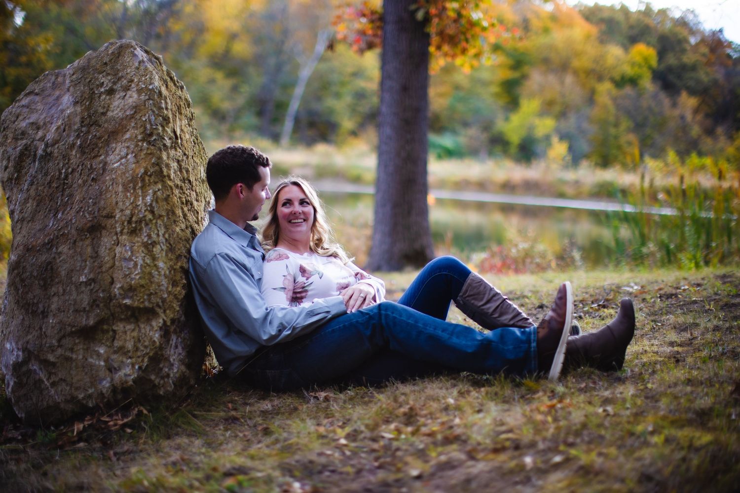 A couple relaxes against a large boulder in an autumn forest setting during golden hour.